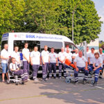 Team des BRK-Patientenfahrdienst Bayreuth mit Spezial- Fahrzeug im Hintergrund und Fahrtrage und Tragestuhl.