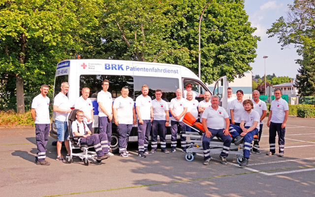 Team des BRK-Patientenfahrdienst Bayreuth mit Spezial- Fahrzeug im Hintergrund und Fahrtrage und Tragestuhl.