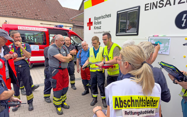 Einsatzkräfte von Rettungsorganisationen, Polizei und Feuerwehr beim Briefing am Einsatzleitwagen des BRK Kreisverbandes Bayreuth. Im Vordergrund befindet sich die sogenannte Fachberaterin Rettungshunde.