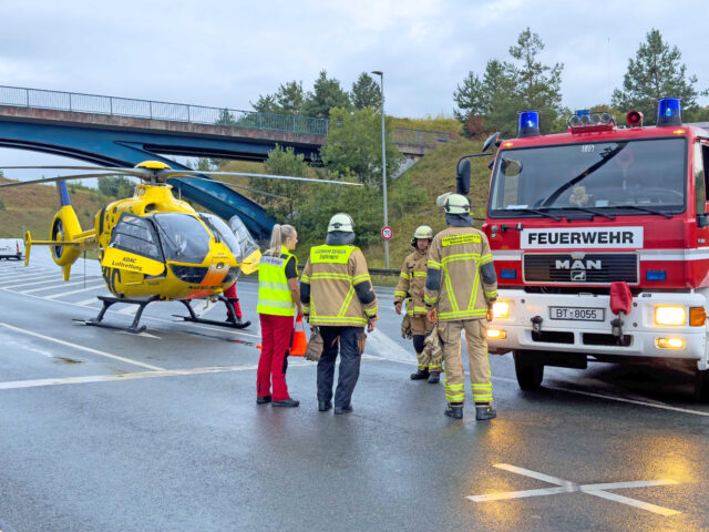 Das BIld zeigt Einsatzkräfte des BRK-Rettungsdienstes Bayreuth, der Feuerwehren und der ADAC-Luftrettung an der Unfallstelle. Im Hintergrund steht ein Rettungshubschrauber der ADAC-Luftrettung und ein Einsatzfahrzeug der Feuerwehren.