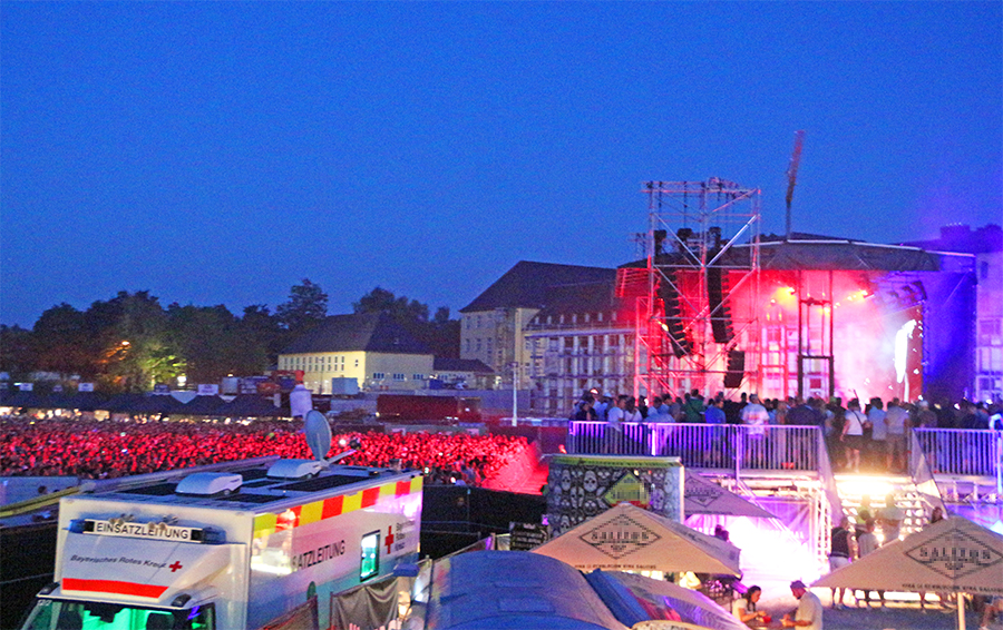 Im Bild die Hauptbühne des Kalkbrenner-Open-Air-Konzertes in Bayreuth bei beginneder Dunkelheit. Sichtbar ist eine Teil der Besucherinnen und Besucher. Im Vordergrund der Leitwagen der BRK-Einsatzkräfte.