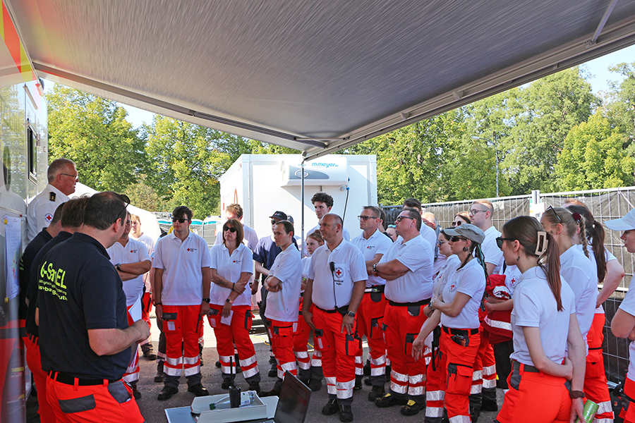 Bild zeigt einen Teil der 55 zur Absicherung des Kalkbrenner-Open-Air-Konzertes in Bayreuth eingesetzten 55 Ehrenamtlchen Helferinnen und Helfer des BRKs bei einem Briefing am Leitwagen.