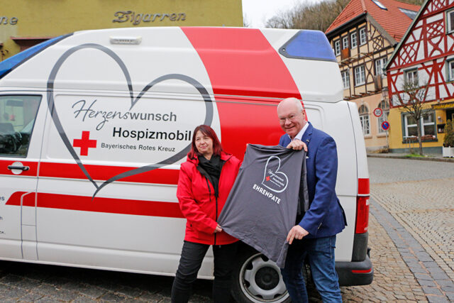 Alexandra Küfner überreicht Martin Rassau ein Shirt mit der Aufschrift Ehrenpate. Im Hintergrund das BRK-Hospizmobil Bayreuth und die Bad Bernecker Altstadt.