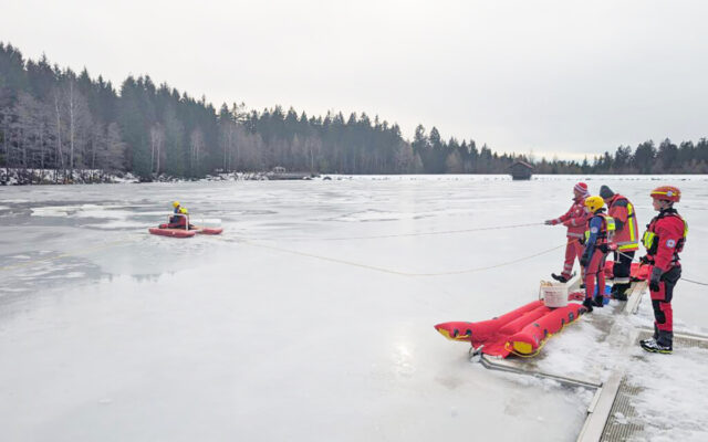 Zu sehen sind Helferinnen und Helfer der Wasserwacht Ortsgruppe Bayreuth bei der Eisrettungsübung am Fichtelsee. Während einige Helferinnen und Helfer vom Steg aus die Retter absichern, arbeiten sich diese mit dem Eisrettungsschlitten bis zur Einbruchstelle vor.