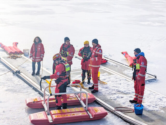 Zu sehen sind Helferinnen und Helfer der Wasserwacht Ortsgruppe Bayreuth bei der Eisrettungsübung am Fichtelsee. Während einige Helferinnen und Helfer vom Steg aus die Retter absichern, bereiten andere Helfer den Eisrettungsschlitten zum Einsatz vor.